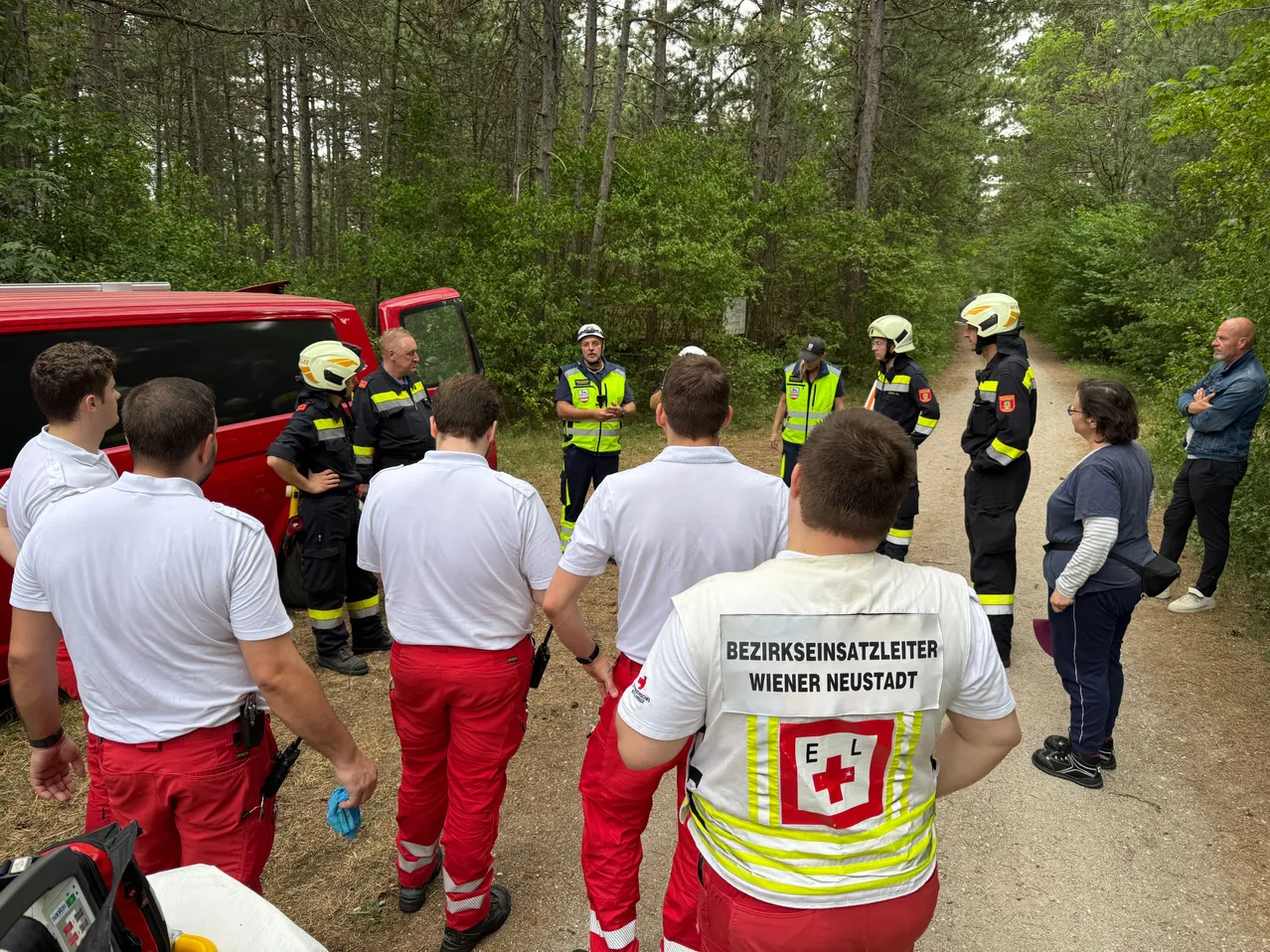 Eine Gruppe von Feuerwehrleuten in roten und gelben Uniformen versammelt sich in einem Waldgebiet und bespricht etwas. Eine Frau in einer blauen Jacke steht auf der rechten Seite.