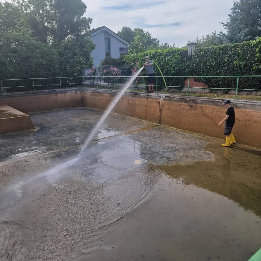Zwei Männer füllen einen großen Pool mit Wasser. Einer hält einen Schlauch und der andere steht an der Seite. Bäume und ein Haus sind im Hintergrund.