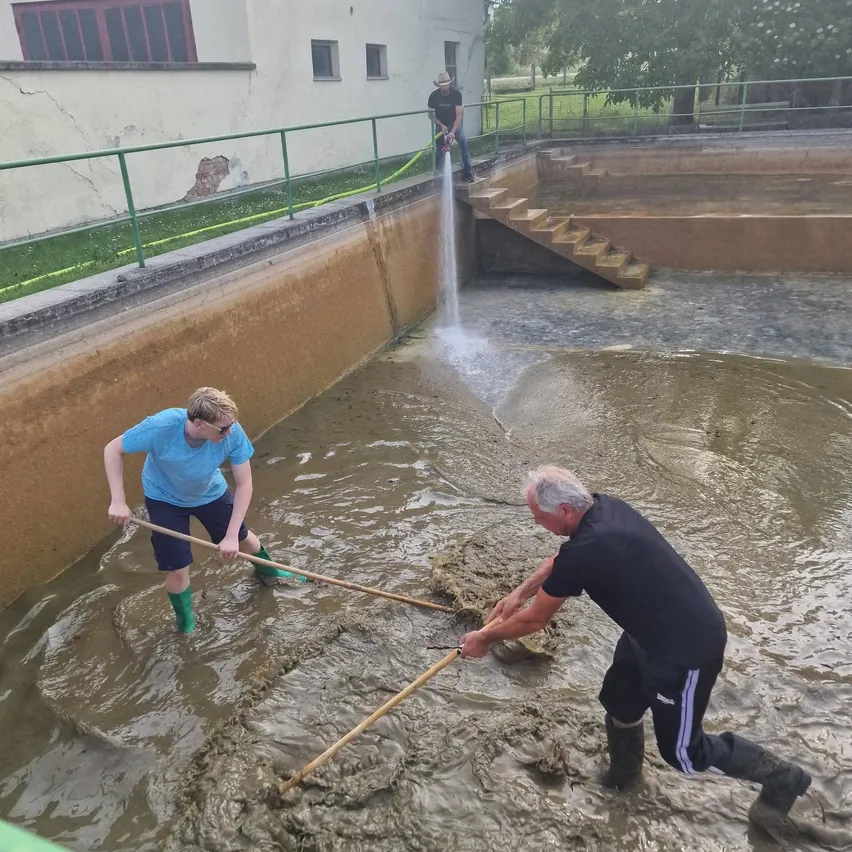 Zwei Personen bewegen mit langen Holzstangen Material durch einen großen schlammigen Teich. Ein Mann steht hinter dem Geländer und beobachtet.