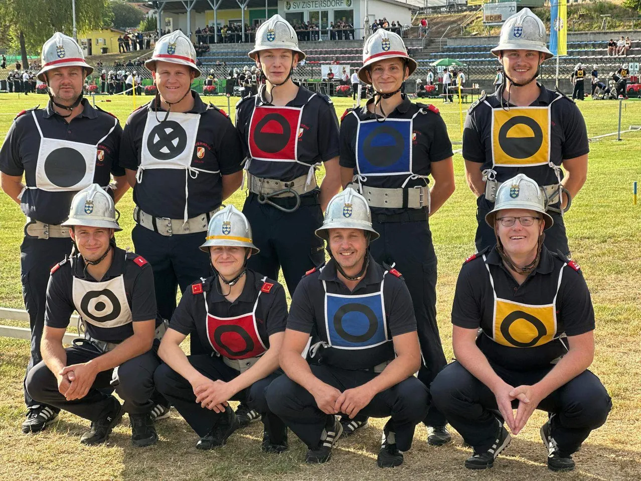 Eine Gruppe von Feuerwehrleuten mit Helmen, Uniformen und Brillen posiert für ein Foto auf einem Rasenfeld.