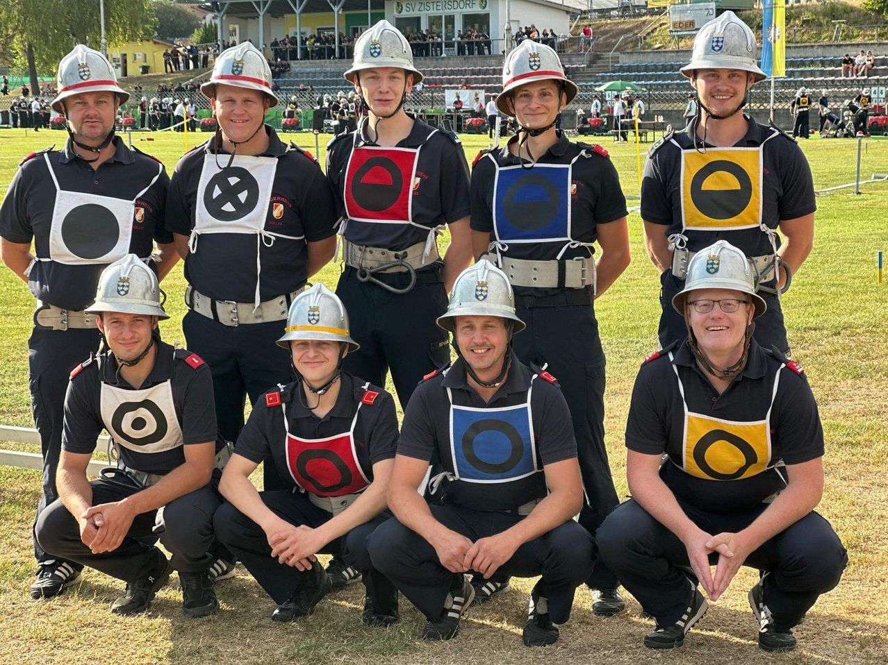 Eine Gruppe von Feuerwehrleuten mit Helmen, Uniformen und Brillen posiert für ein Foto auf einem Rasenfeld.