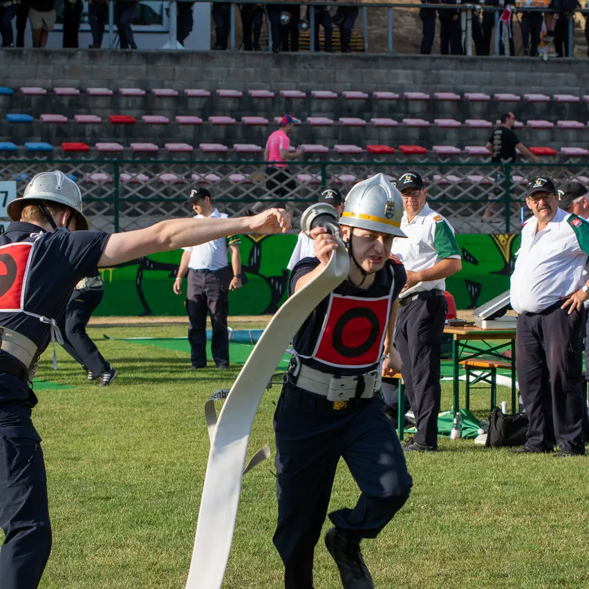 Mehrere Personen laufen auf einem Grasfeld in Sicherheitsausrüstung. Einer von ihnen hält ein aufgerolltes Objekt. Dahinter stehen Menschen, die zuschauen, und einige sitzen auf Tribünen.