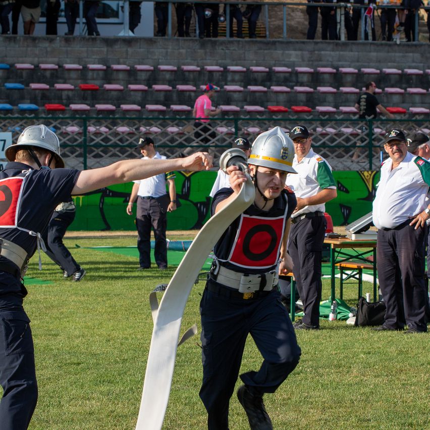 Mehrere Personen laufen auf einem Grasfeld in Sicherheitsausrüstung. Einer von ihnen hält ein aufgerolltes Objekt. Dahinter stehen Menschen, die zuschauen, und einige sitzen auf Tribünen.