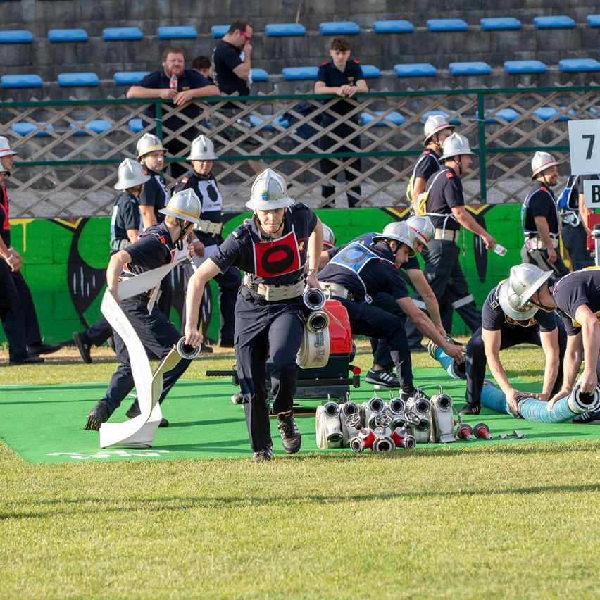 Eine Gruppe von Feuerwehrleuten läuft auf einem grünen Feld und hält Feuerlöscher und Schläuche in der Hand. Sie tragen Helme und Uniformen. Zuschauer stehen im Hintergrund und beobachten sie. Dahinter befindet sich eine grüne Wand mit Graffiti.