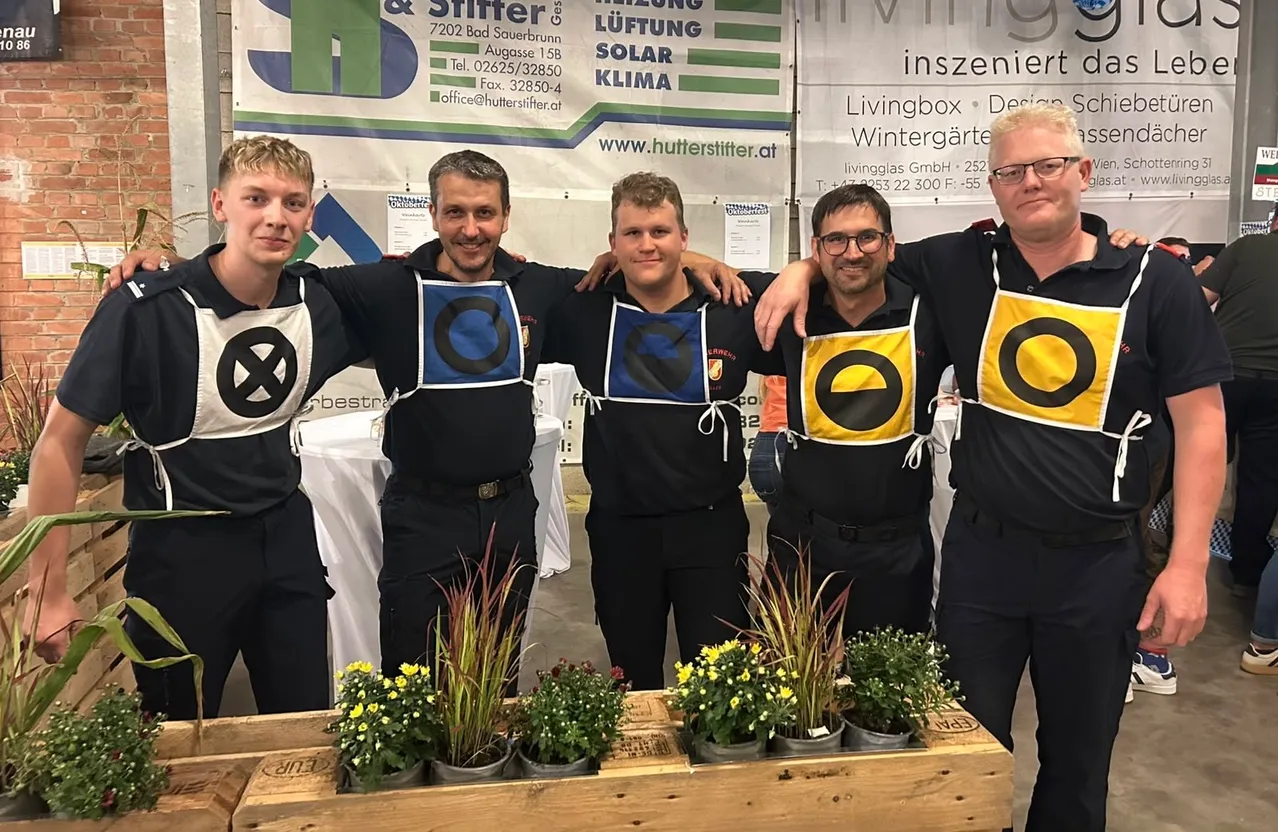Fünf Männer in gleichen Uniformen stehen hinter einem Holztisch mit Topfpflanzen und lächeln für ein Foto. Dahinter ist ein Banner mit Text und Logos.