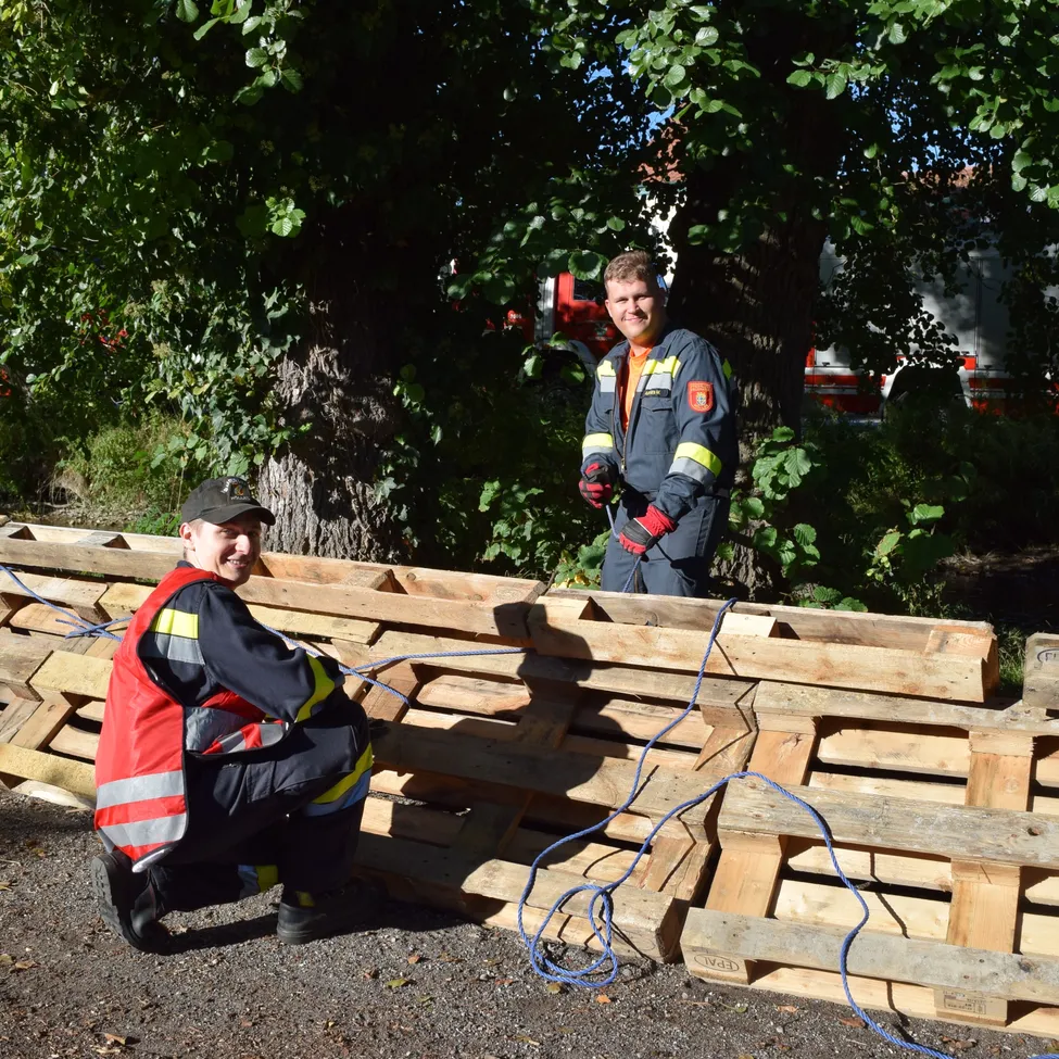 Zwei Feuerwehrleute arbeiten an einer Holzbarriere mit blauen Kabeln im sonnigen Freien.