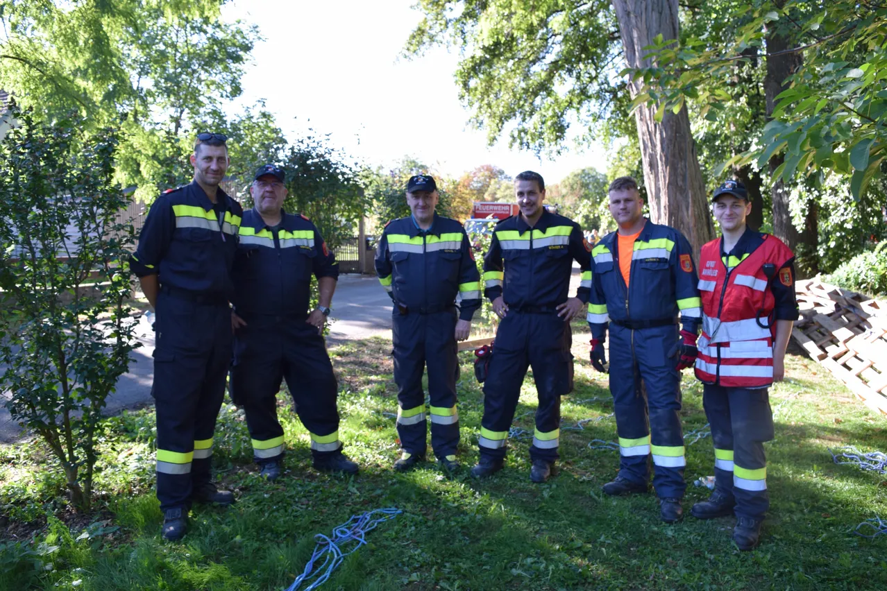 Fünf Feuerwehrleute in Uniform stehen in einem Park und posieren für ein Foto. Sie lächeln und scheinen eine Pause einzulegen.