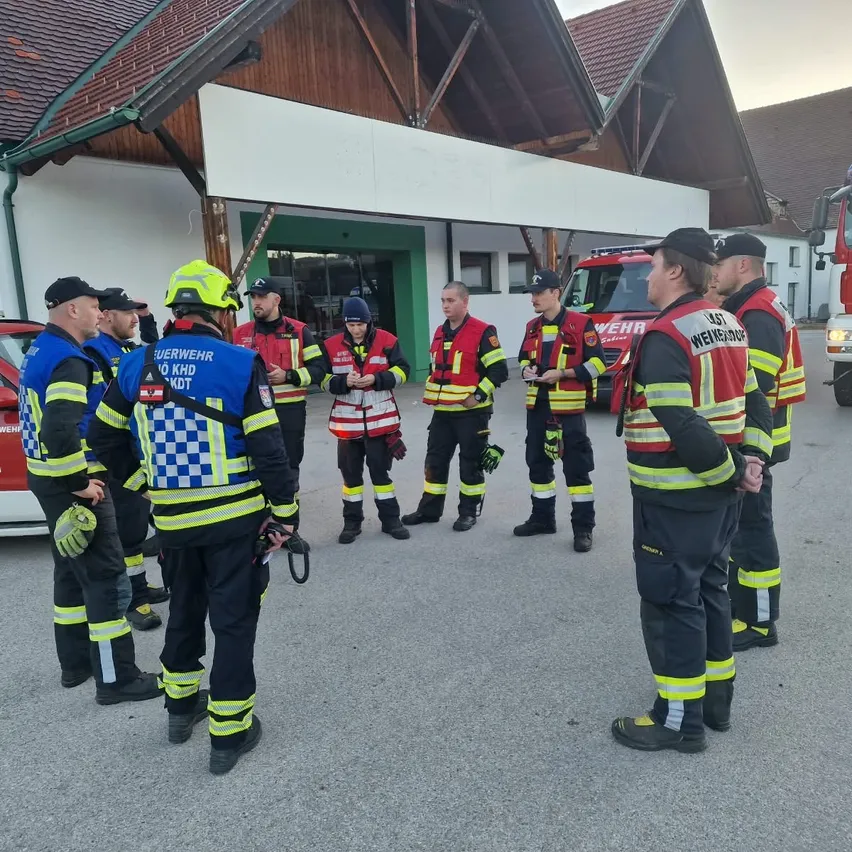 Eine Gruppe von Feuerwehrleuten in Uniform steht vor einem Feuerwehrhaus und bespricht etwas.