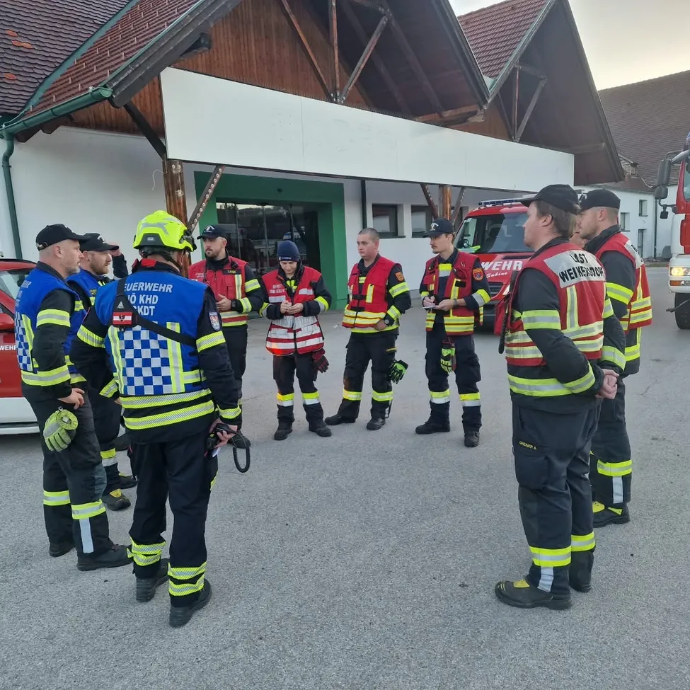 Eine Gruppe von Feuerwehrleuten in Uniform steht vor einem Feuerwehrhaus und bespricht etwas.