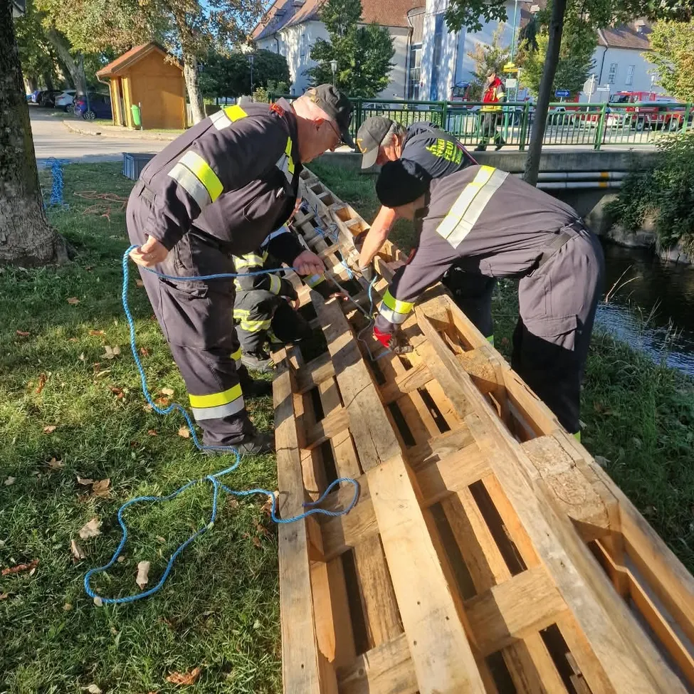 Zwei Feuerwehrleute in Uniform arbeiten an einem Holzpalette auf einer Rasenfläche in der Nähe eines Flusses.