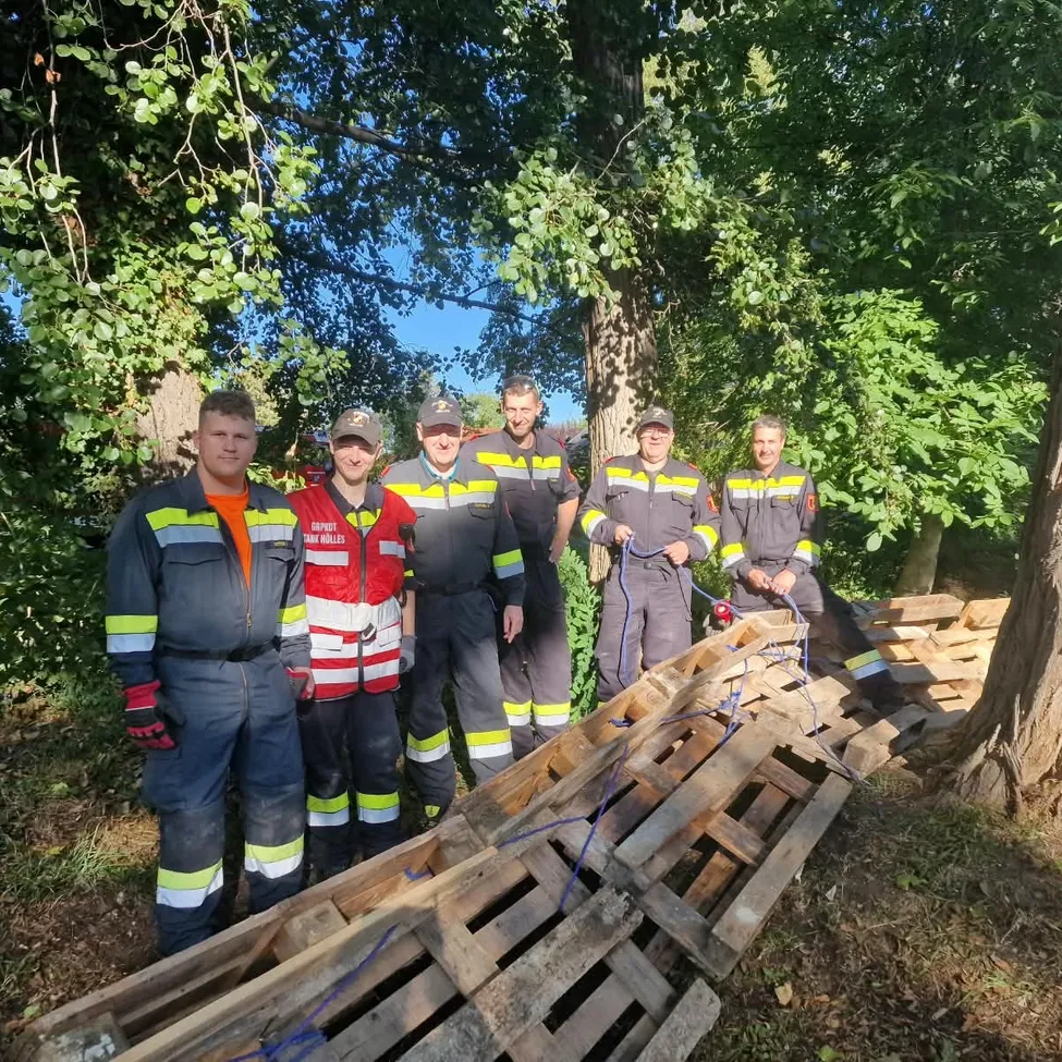 Eine Gruppe von Feuerwehrleuten in Uniform steht neben einem Haufen Holzpaletten im Wald. Sie lächeln und scheinen für ein Foto zu posieren.