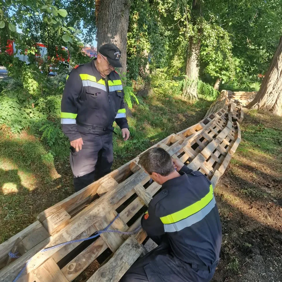 Zwei Feuerwehrleute in Uniform arbeiten an einer Holzstruktur in einem Park. Einer kniet und repariert etwas, während der andere steht und zusieht.
