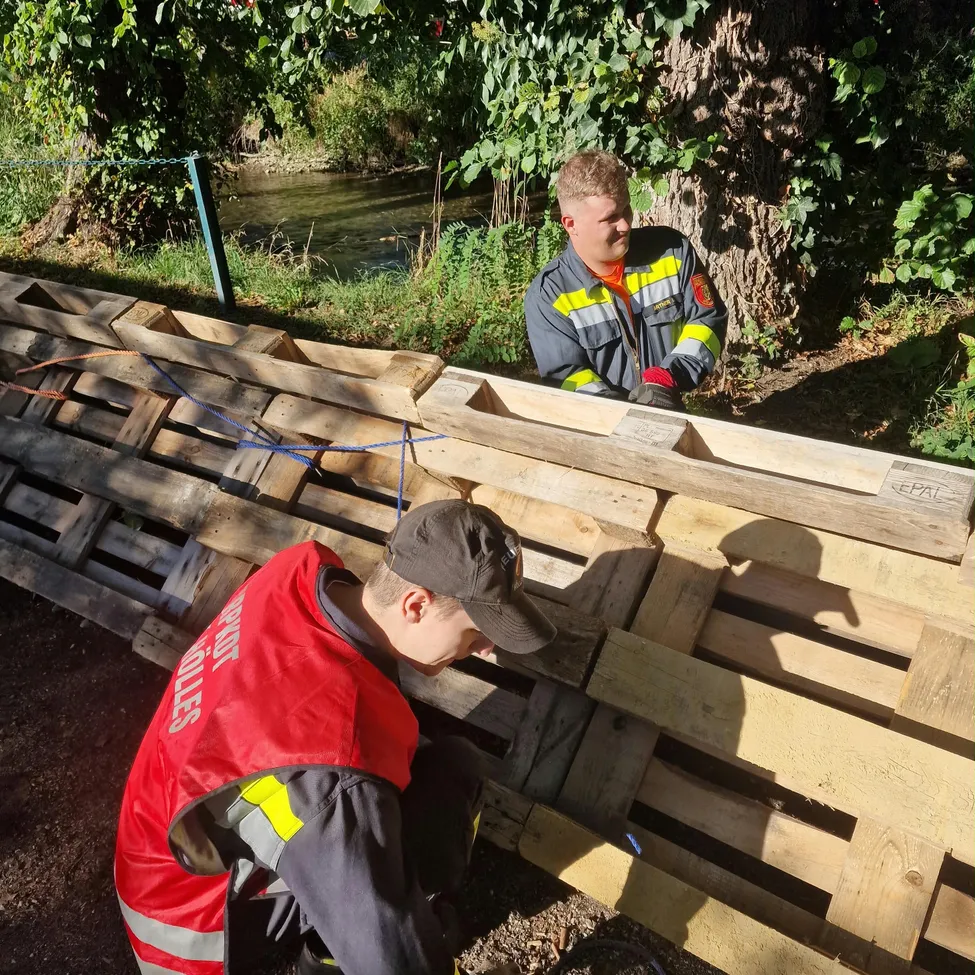Zwei Feuerwehrleute in Uniform arbeiten an einer Holzstruktur in der Nähe eines Flusses, einer kniet und der andere steht hinter ihm.