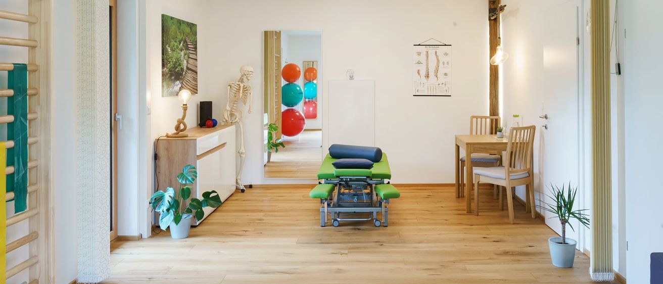 Ein leerer Raum mit Holzböden, weißen Wänden und Deckenbeleuchtung. Ein Skelettmodell ist auf der linken Seite, ein Tisch mit Stühlen auf der rechten Seite und ein Massagetisch in der Mitte. Ein Spiegel reflektiert den Raum.