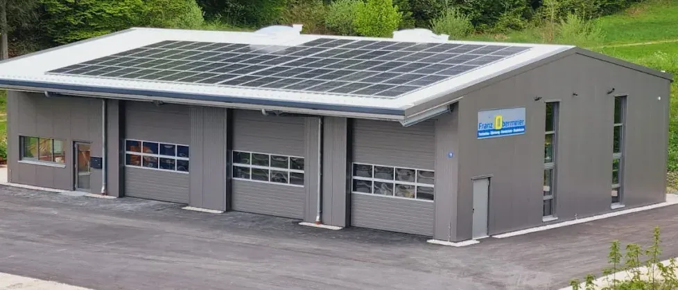 Ein graues Gebäude mit einem Solardach und einem Schild mit der Aufschrift Franz Berner. Das Gebäude hat mehrere Fenster und Türen und ist von Bäumen umgeben.