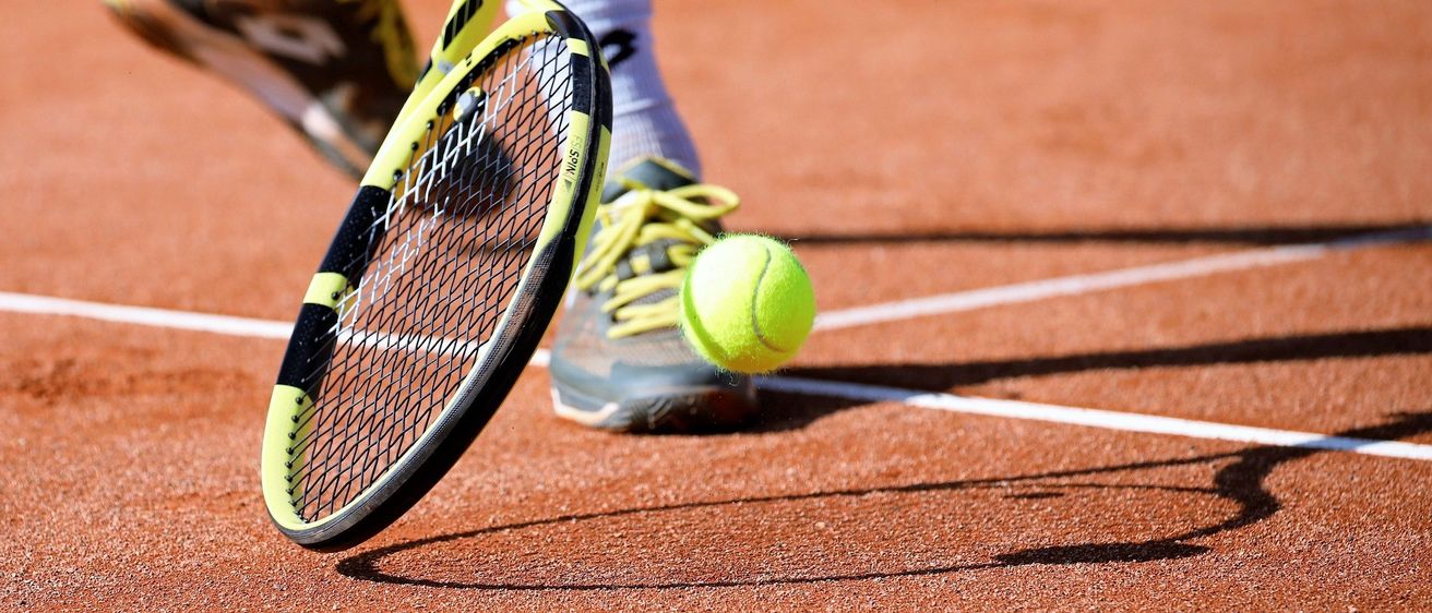 Der Fuß und der Schläger eines Tennisspielers sind im Fokus, mit einem Tennisball auf der Oberfläche des Platzes.