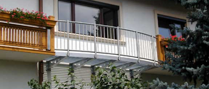 Ein weißes Gebäude mit einem Balkon, der mit Metallgeländern und Blumenkästen ausgestattet ist. Zwei Fenster und ein Schornstein sind am Gebäude zu sehen.