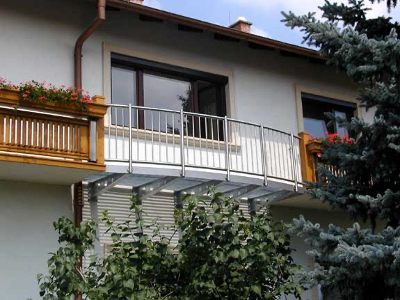 Ein weißes Gebäude mit einem Balkon, der mit Metallgeländern und Blumenkästen ausgestattet ist. Zwei Fenster und ein Schornstein sind am Gebäude zu sehen.