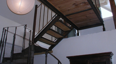 Eine Wendeltreppe mit Holzstufen und Metallgeländern. Sie führt von einer weiß gestrichenen Wand zu einer Holzdecke.