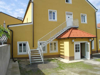 Ein strahlend gelbes Haus mit weißen Fensterrahmen, ausgestattet mit einer Metalltreppe zum Balkon und einer offenen Tür.