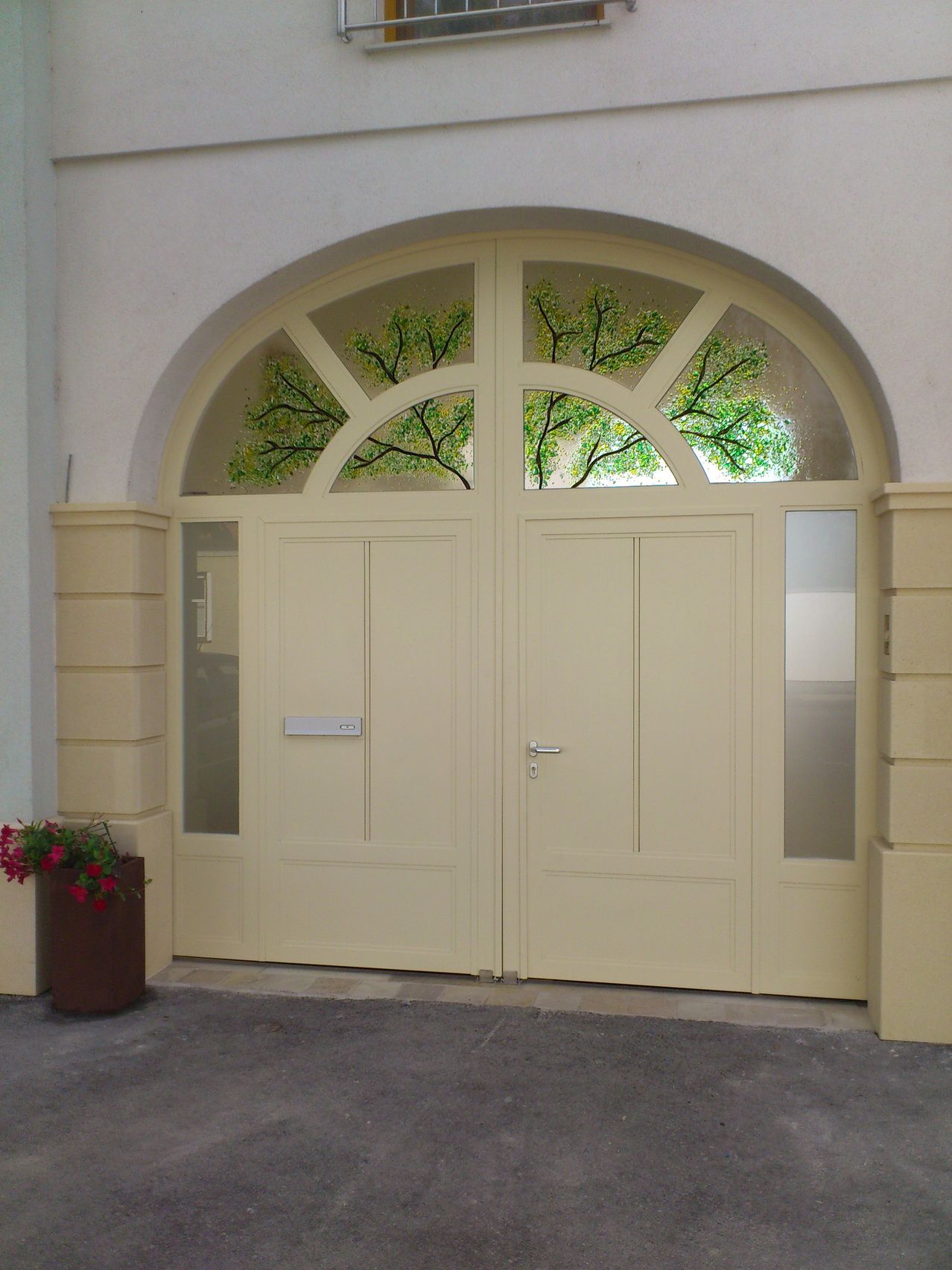Eine beige Doppeltür mit einem großen Bogenfenster darüber. Das Fenster zeigt ein Buntglasdesign eines Baumes. Ein Topf mit roten Blumen steht links neben der Tür.