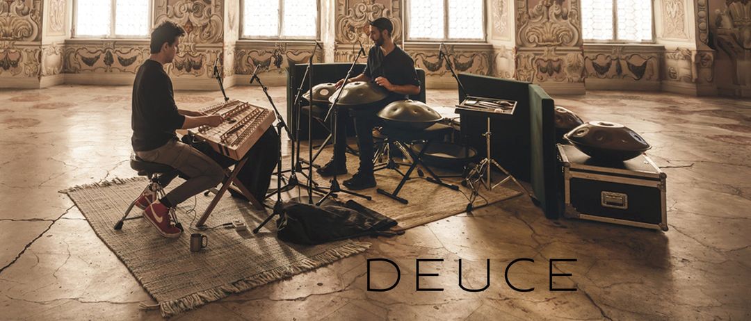 Zwei Männer spielen Instrumente in einem großen Saal mit verzierten Wänden und bogenförmigen Fenstern. Einer spielt Hackbrett, der andere Gong. Der Text 'MANU DELAGO & MAX ZT' und 'DEUCE' ist auf dem Bild zu sehen.