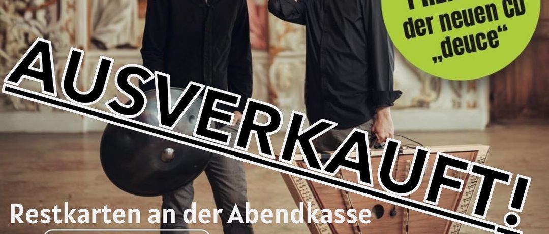 Zwei Männer stehen vor einer Wand mit einem Bild eines Heiligen. Der Mann auf der linken Seite hält eine große Trommel. Der Text an der Wand lautet 'AUSVERKAUFT!' und 'Welt-Premiere der neuen CD'. Die Veranstaltung findet im Theater Garage Sulz, Österreich am 12.3.2026 um 20:30 statt.