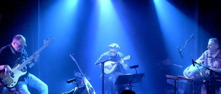 Ein Musiker spielt eine Laute auf einer Bühne unter blauer Bühnenbeleuchtung. Ein anderer Musiker spielt Gitarre hinter ihm, mit anderen Instrumenten in der Nähe. Das Publikum sieht von den Schatten zu.