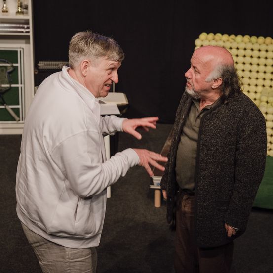 Drei Männer unterhalten sich auf einer Bühne. Einer trägt eine weiße Jacke, der andere eine graue. Dahinter befindet sich ein großes, gelbes Objekt, das einer Balldisplay ähnelt.