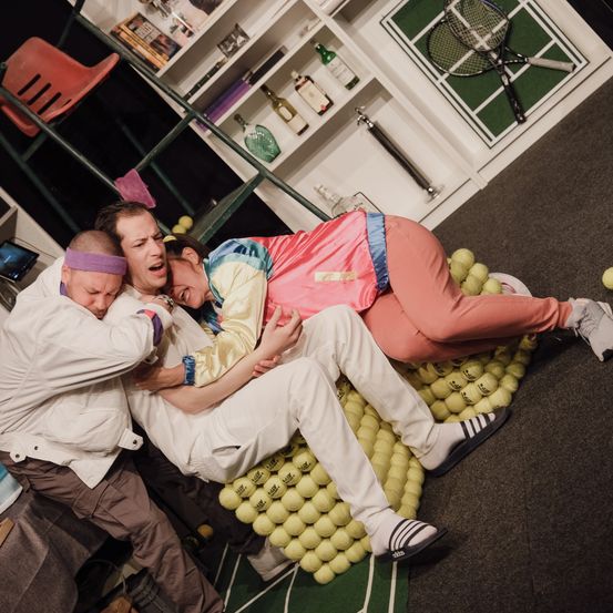 Drei Personen sind auf einer Bühne, eine davon liegt auf einem Haufen Tennisbälle. Sie tragen farbenfrohe Kleidung und scheinen in einem Theaterstück zu agieren.