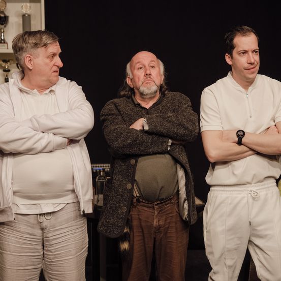 Drei Männer stehen nebeneinander mit verschränkten Armen. Der Mann auf der linken Seite trägt eine weiße Kapuzenjacke. Der Mann in der Mitte hat graues Haar und trägt eine braune Jacke. Der Mann auf der rechten Seite trägt ein weißes Hemd und eine Uhr. Hinter ihnen befindet sich ein dunkler Hintergrund und einige gelbe Objekte.