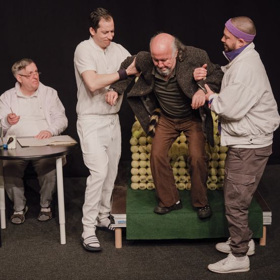 Drei Männer in weißen Kitteln und einer in einem lila Stirnband unterstützen einen älteren Mann in einem grauen Mantel und braunen Hosen. Sie stehen auf einer Bühne mit einem Tennisballhintergrund.