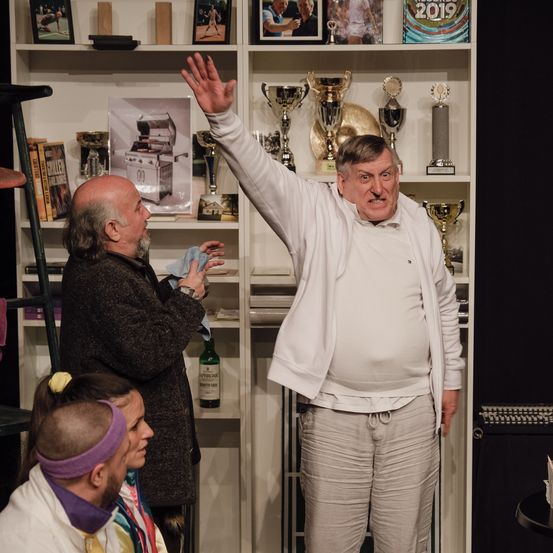 Mehrere Personen versammeln sich in einem Raum; ein Mann in einer weißen Jacke winkt, während ein anderer in einem schwarzen Mantel ein Tuch hält. Hinter ihnen sind Pokale auf Regalen ausgestellt.