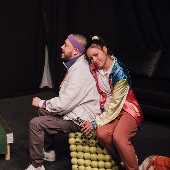 Ein Mann sitzt auf einem Tennisballstuhl und sieht ernst aus, während ein Paar auf einem anderen Tennisballstuhl sitzt und ihn ansieht, wobei eine Frau eine Flasche in der Hand hält.