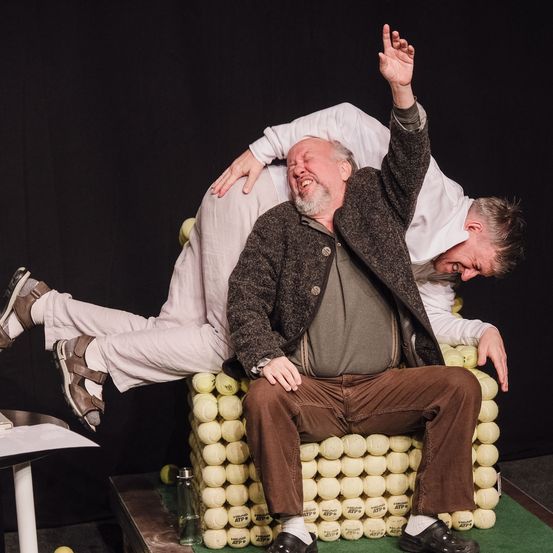 Zwei Männer auf der Bühne, einer sitzt auf einem Stuhl aus Tennisbällen und der andere kniet hinter ihm, beide lächeln.
