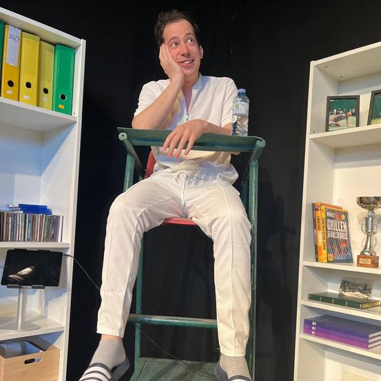 Ein Mann sitzt auf einem grünen Stuhl, trägt weiße Hosen und ein weißes Hemd, lächelt und posiert für ein Foto. Hinter ihm befinden sich Regale mit Büchern, Trophäen und Ordnern. Eine Wasserflasche steht auf dem Stuhl.