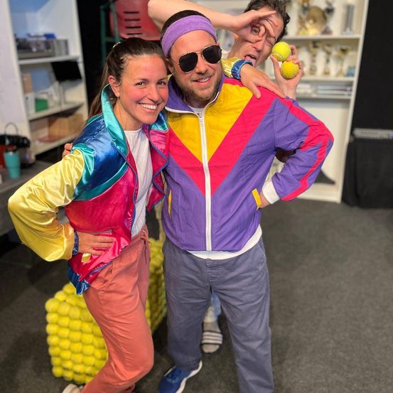 Ein lächelndes Paar in Retro-Sportkleidung steht neben einer Person, die Tennisbälle hält. Sie befinden sich in einem Raum mit einem Haufen gelber Tennisbälle und Regalen im Hintergrund.