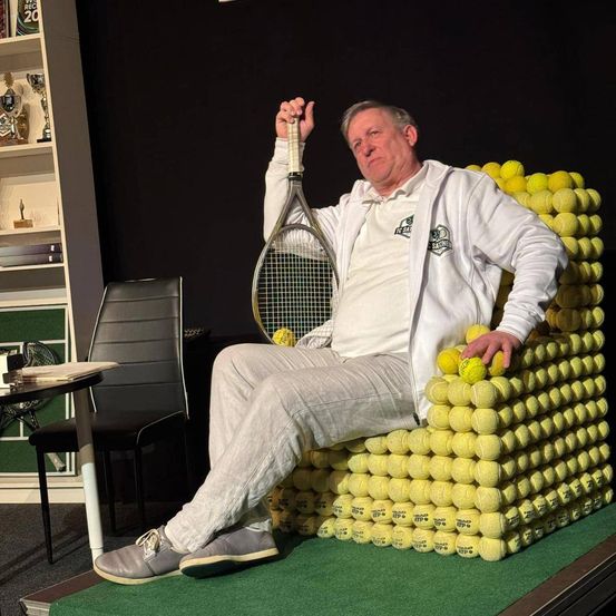 Ein Mann sitzt auf einem Stuhl aus Tennisbällen, hält einen Tennisschläger und schaut nach rechts. Hinter ihm steht ein schwarzer Stuhl und auf der linken Seite ein Schreibtisch mit einem Buch. Im Hintergrund befinden sich Regale mit Trophäen und Büchern.