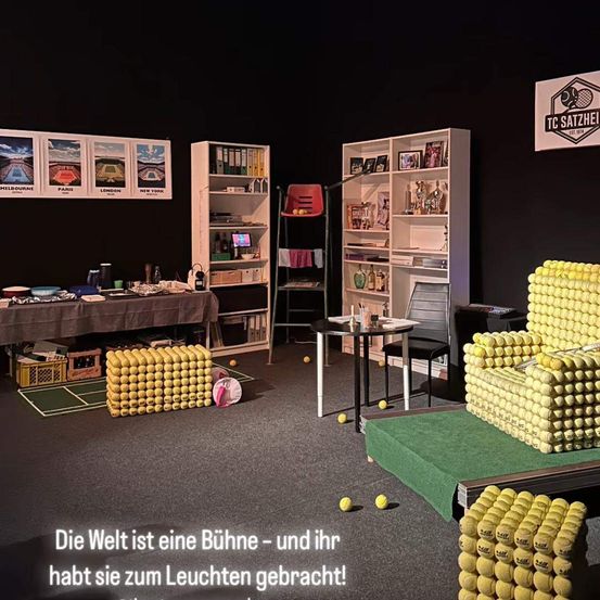 Ein Raum mit verschiedenen kreativ angeordneten Tennisbällen, einschließlich eines Stuhls, eines Tisches und Regale. Der Raum ist mit Bildern von Tennisplätzen dekoriert. Der Satz 'Die Welt ist eine Bühne - und ihr habt sie zum Leuchten gebracht!' ist auf Deutsch geschrieben.