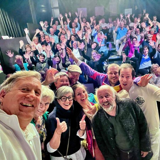 Eine Gruppe von Menschen, die feiern, einige mit erhobenen Händen, in einem Theater. Ein Mann vorne lächelt, während andere klatschen. Der Hintergrund zeigt eine Menge mit unterschiedlichen Ausdrücken.