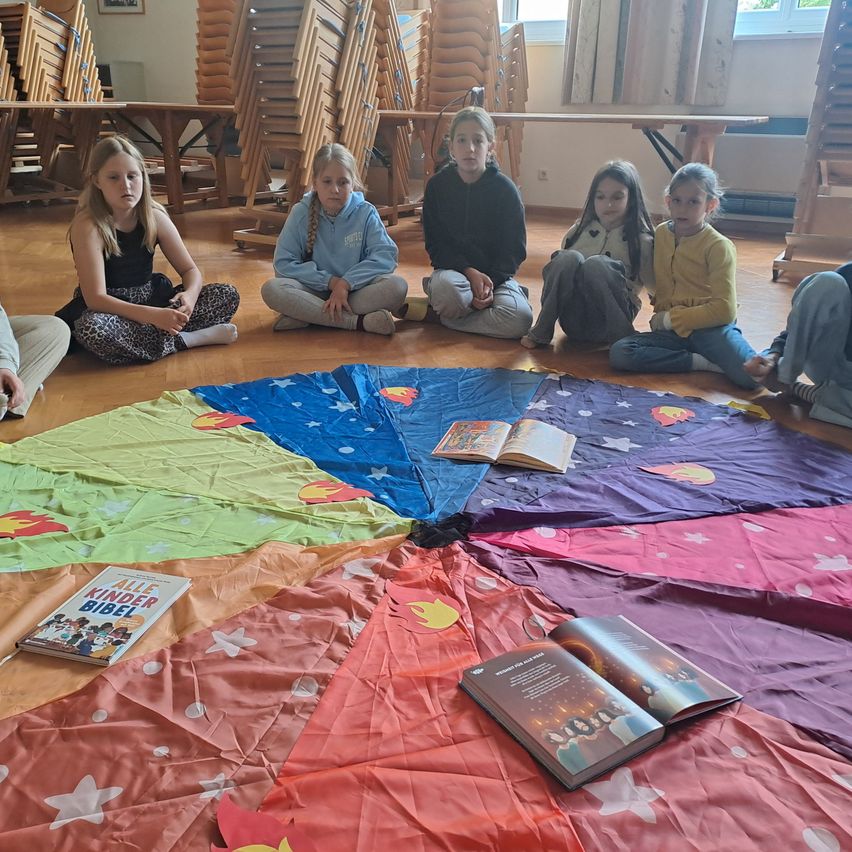 A group of young girls sit around a colorful circle on the floor. They are reading books and looking at the camera. Behind them are wooden chairs and a wooden table.