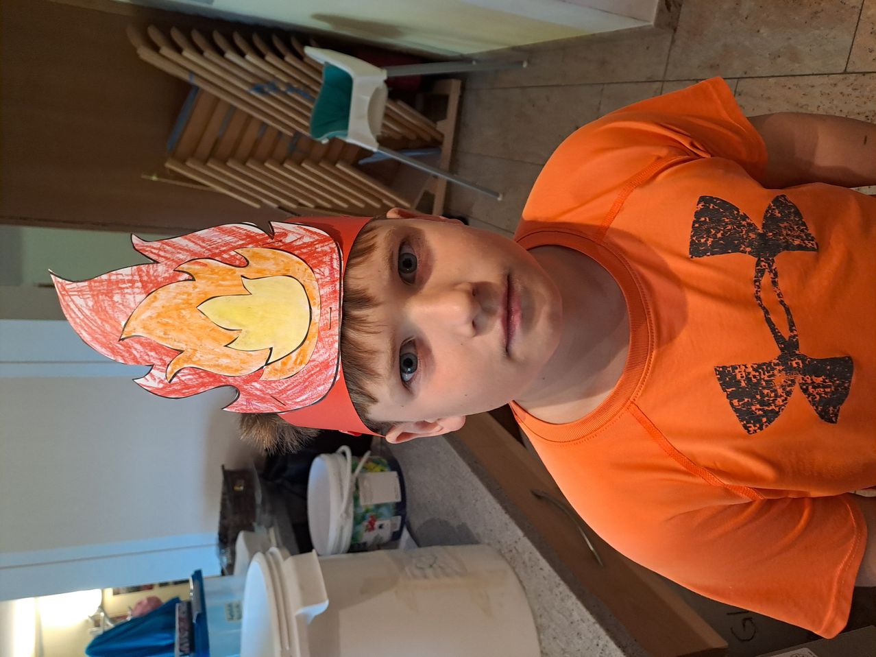 A young boy wearing an orange shirt and a fireman's hat with flames looks straight ahead. Behind him, chairs are stacked, and various items are on the floor.