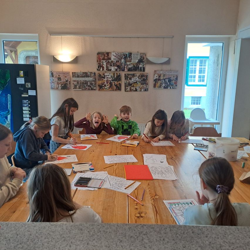 A group of children sit at a long table, coloring in pages with various art supplies on the table.