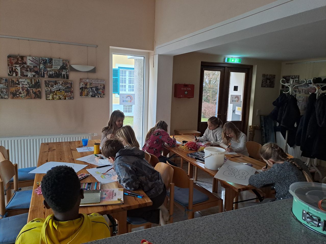 A group of children and adults are gathered around tables in a room, engaged in an art activity. The tables are covered with papers, coloring materials, and a white bucket. A door leads to a room with windows and a sign.