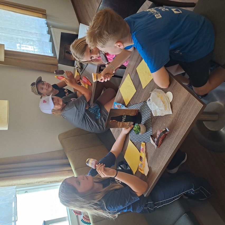 Eine Gruppe von Kindern sitzt an einem Tisch in einem Hotelzimmer und isst Eis und Snacks. Ein Mädchen in einem blauen Shirt hält eine kleine Pflanze.