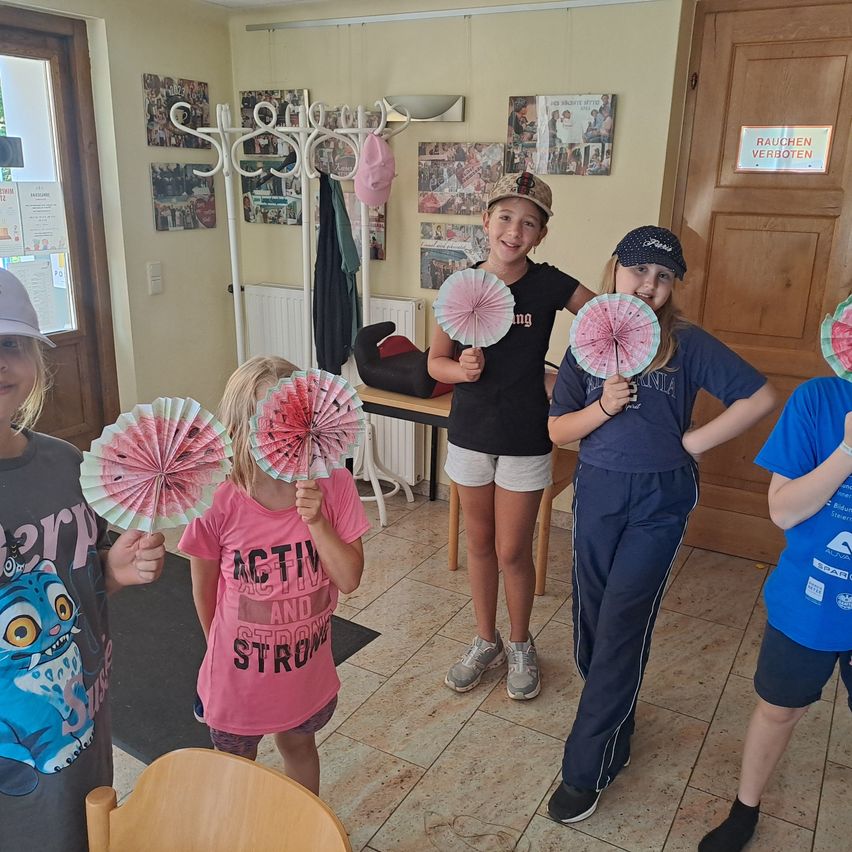 Eine Gruppe von Kindern ist in einem Raum und hält rosa Papierfächer. Sie lächeln und posieren für ein Foto. Der Raum hat gefliesten Boden, einen weißen Heizkörper und eine Tür mit einem Schild, das 'Bauchen Verboten' sagt.