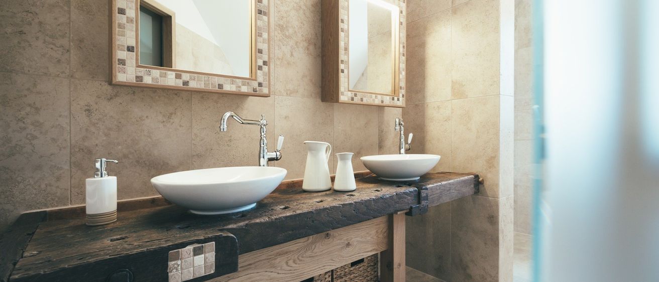 Ein Badezimmer mit zwei Waschbecken und Armaturen, gefliesten Wänden, Holzarbeitsplatten und zwei Spiegeln. Ein Seifenspender befindet sich links.