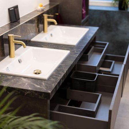 Ein Badezimmer mit zwei weißen Waschbecken, beide mit goldenen Armaturen, an einer Marmortheke. Die Waschbecken werden von mehreren grauen Schubladen flankiert.