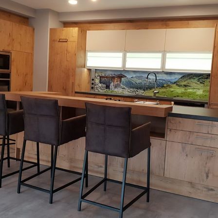 Eine moderne Küche mit Holzschränken und Arbeitsplatten. Zwei braune Stühle sind am Tresen positioniert, mit einer Aussicht auf eine Berglandschaft an der Wand.