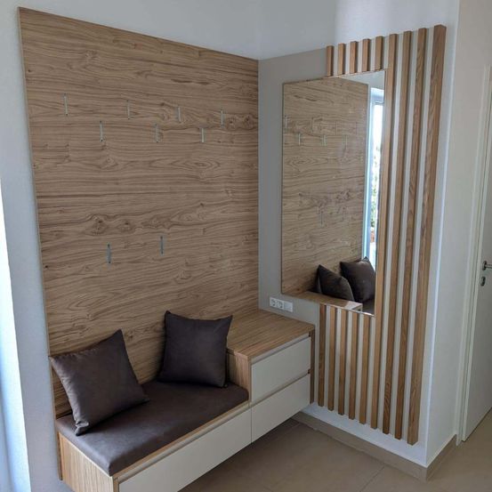 Ein modernes Interieur zeigt eine Holzwand mit Haken, eine Bank mit zwei Kissen und einen großen Spiegel. Der Raum hat eine weiße Wand und einen Holzboden.